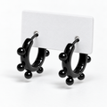 Pair of glossy black stainless steel huggie hoops with round ball studs displayed on a white holder