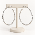 Pair of matte silver-tone wavy post-back hoop earrings displayed on an ivory earring holder