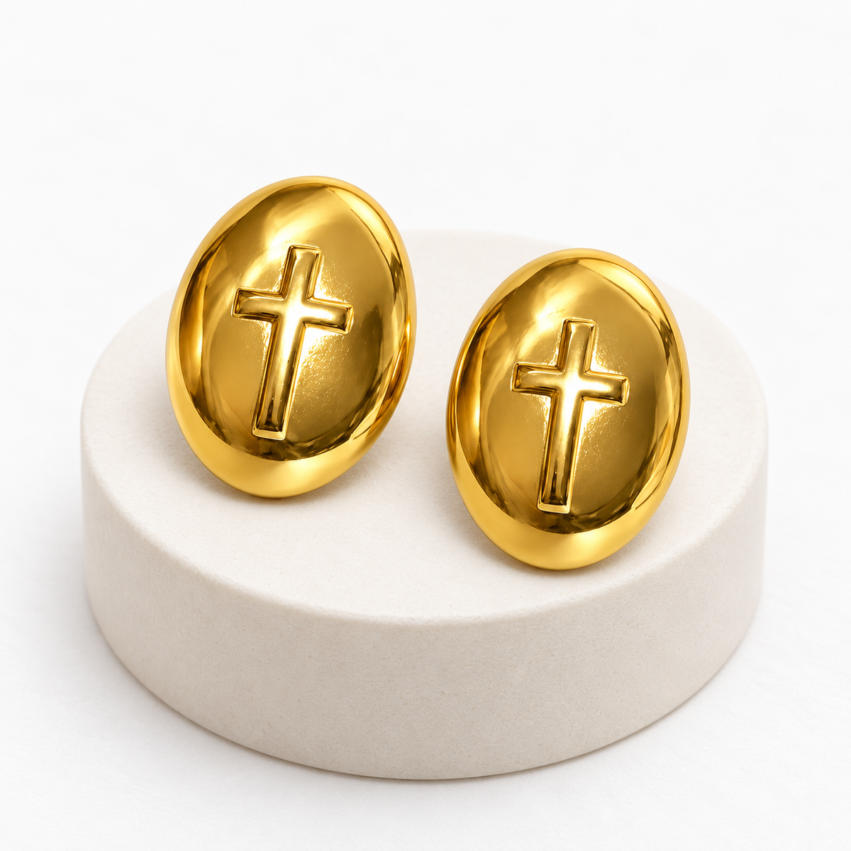 Pair of gold-tone stainless steel oval cross post-back stud earrings resting on an ivory pedestal